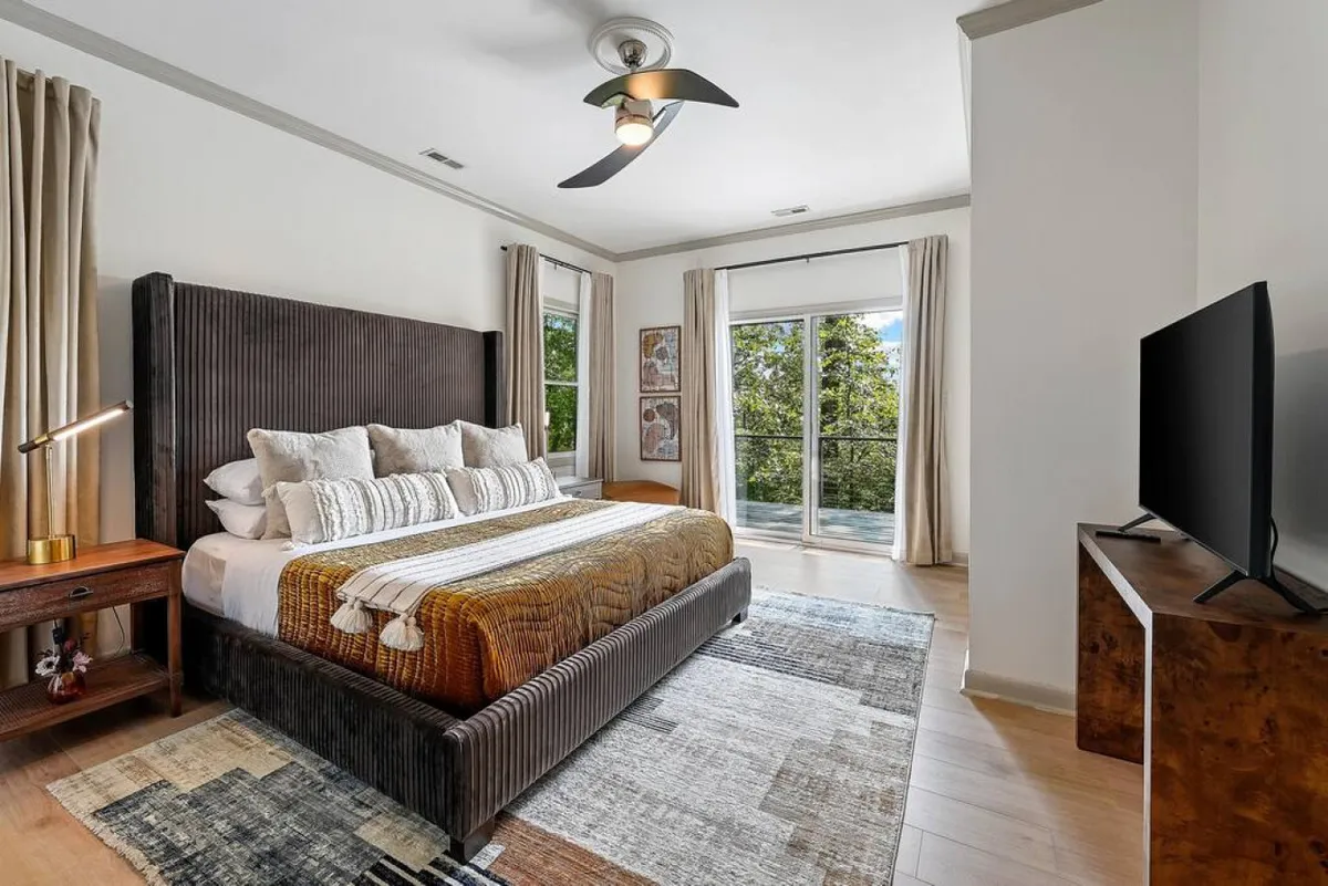 A serene king-size bedroom with a textured headboard, colorful decorative pillows, a large window streaming in natural light, a geometric-patterned rug, and a cozy seating area. The space feels both spacious and welcoming, ideal for restful nights and quiet mornings.