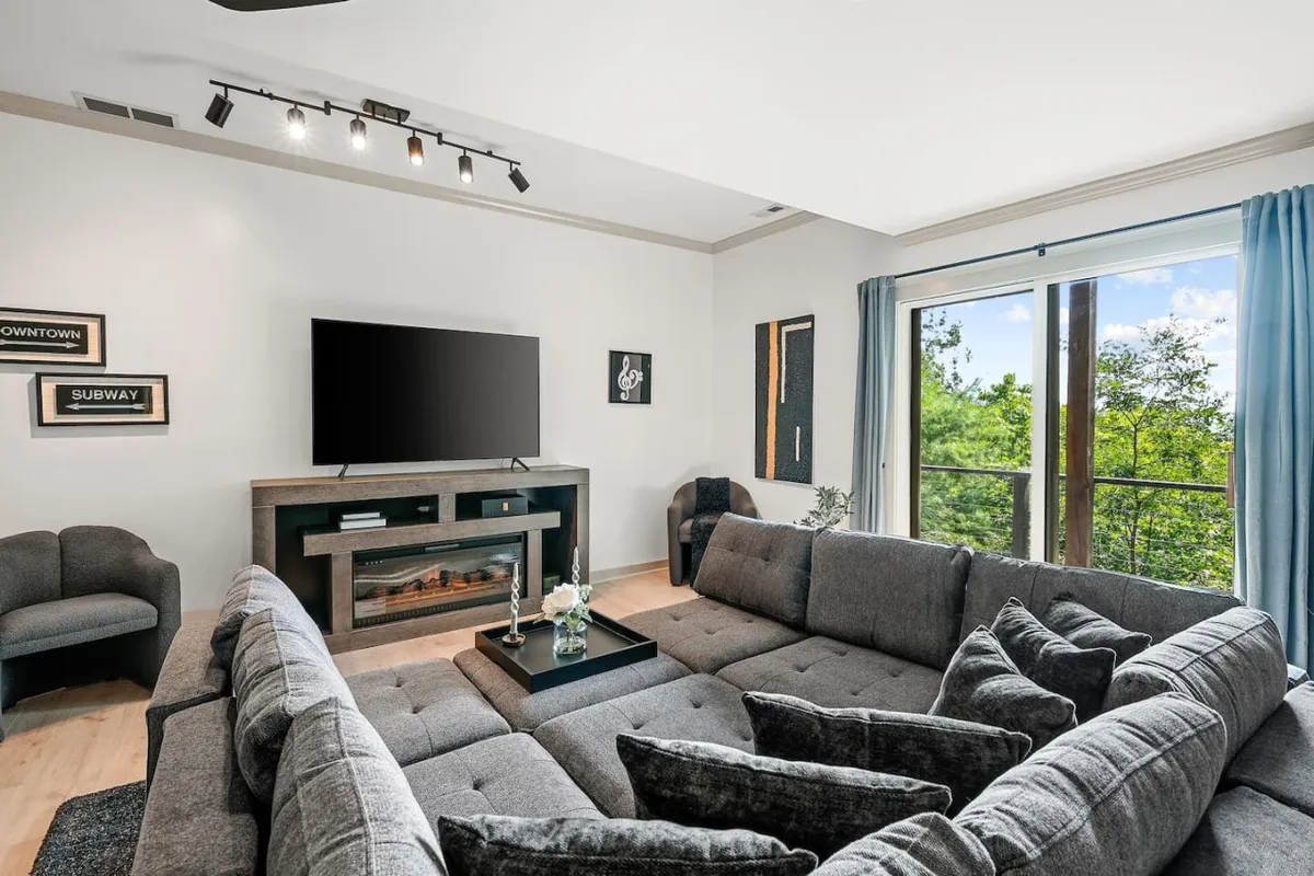 A spacious living area with a large sectional sofa facing a TV unit and fireplace, framed by floor-to-ceiling windows that flood the room with natural light. The arrangement is perfect for group entertainment and relaxation.