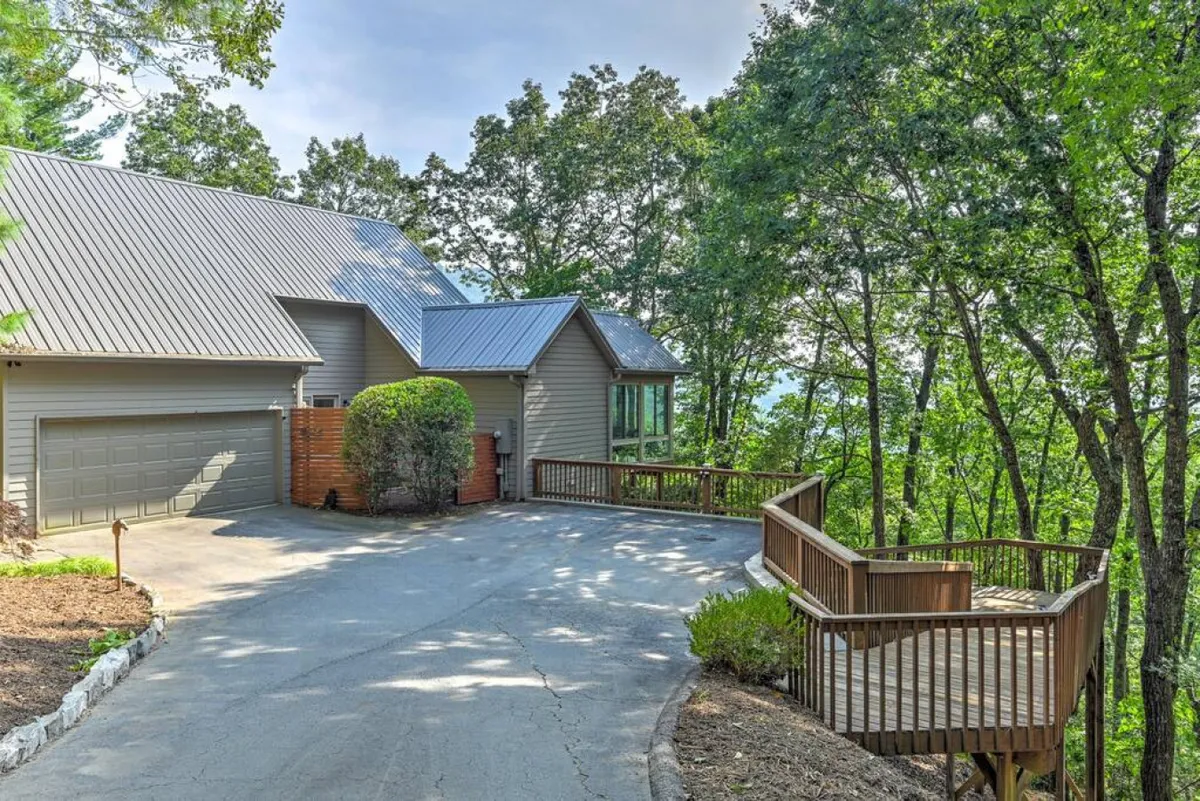 The image features a spacious wooden deck attached to the house, overlooking lush landscape greenery and bordered by a driveway. The house façade is visible, and the deck provides ample space to relax outdoors while enjoying the serene, natural setting.