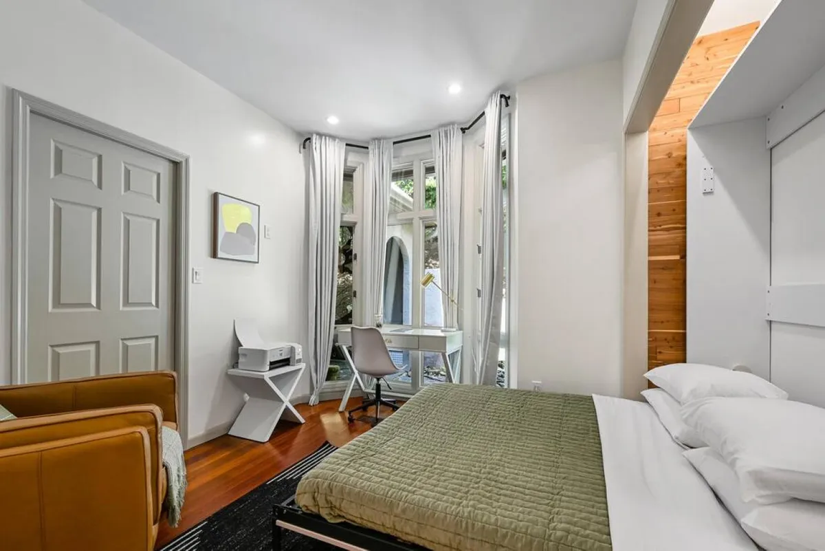 A thoughtfully appointed flexible bedroom/office featuring a comfortable bed with a green quilt, a writing desk and chair by large windows letting in ample natural light. Subtle decor and soft colors make it inviting for both work and relaxation.