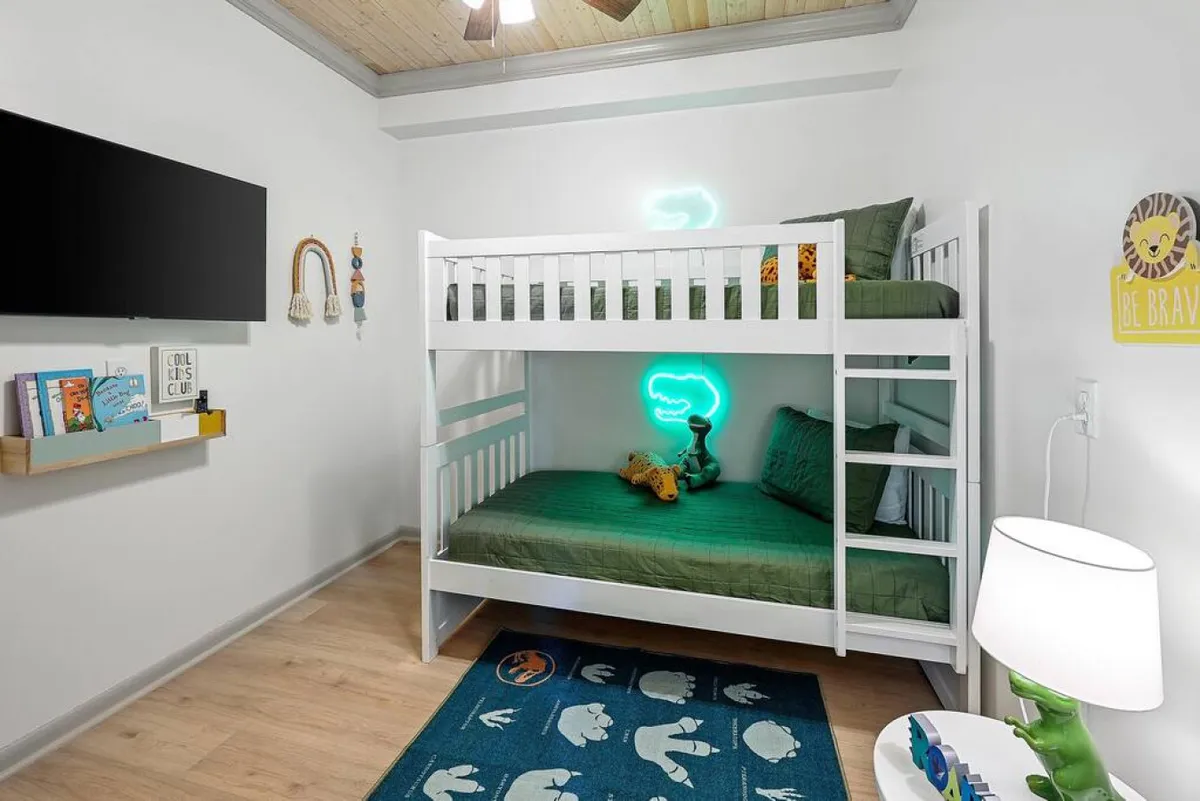 A cheerful bunk room with a built-in bunk bed, playful neon light, bookshelf filled with books, and a cozy rug—creating an inviting space for kids or the young-at-heart. Soft lighting and clever storage add to its charm.