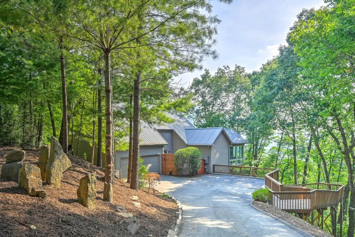 A wide shot of the property exterior shows the driveway leading up to the house, natural stone features, a welcoming deck area, and a lush, wooded setting. The image evokes arrival, anticipation, and the communal excitement of starting a mountain retreat together.