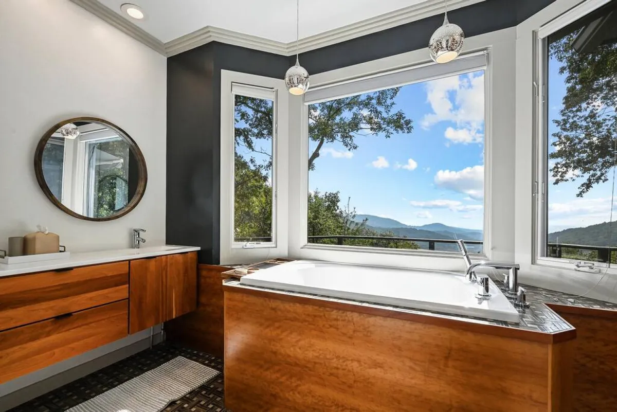 A spa-inspired bathroom complete with a freestanding bathtub, expansive windows offering mountain views, round mirror, and elegant wood cabinetry. The space exudes luxury and relaxation, ideal for self-care and unwinding after a long day.