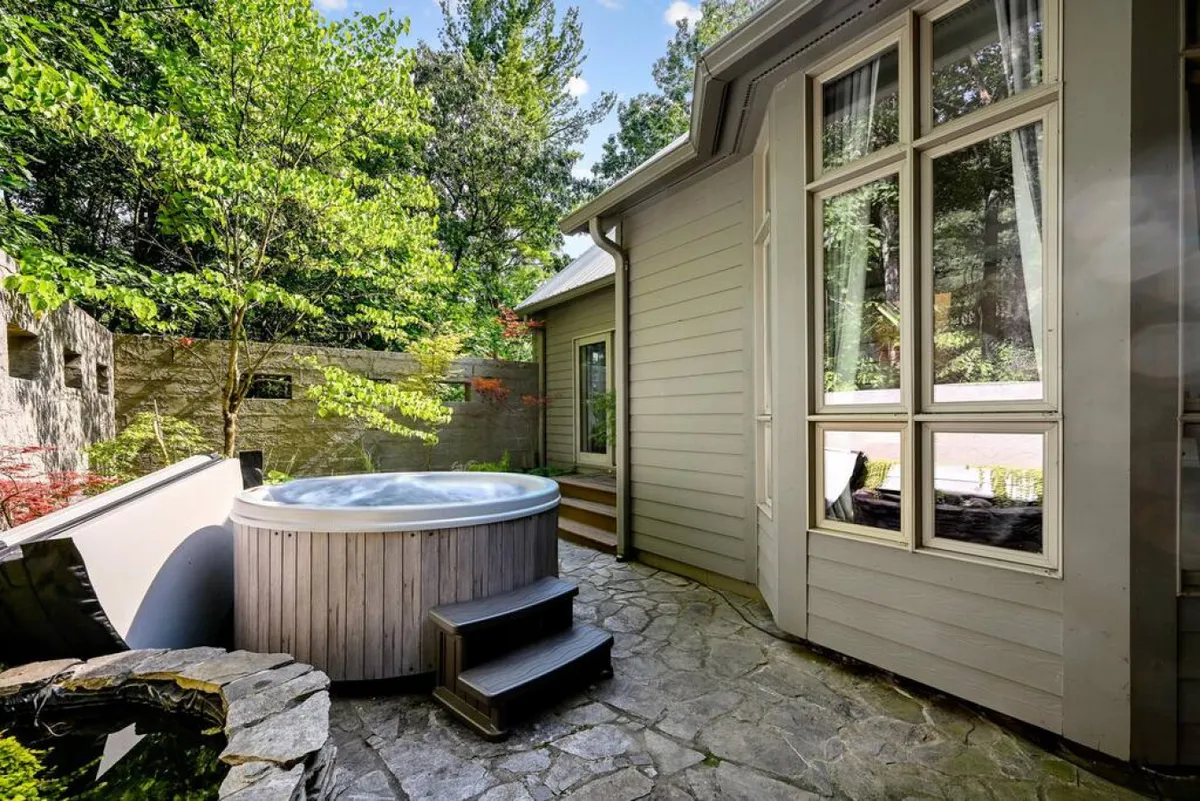 This exterior image features a 6-person hot tub set along a natural stone pathway, surrounded by lush greenery and large windows that hint at the welcoming interior. The scene exudes tranquility and offers an inviting space for guests to relax, socialize, and embrace the mountain landscape at sunset or beneath the stars.