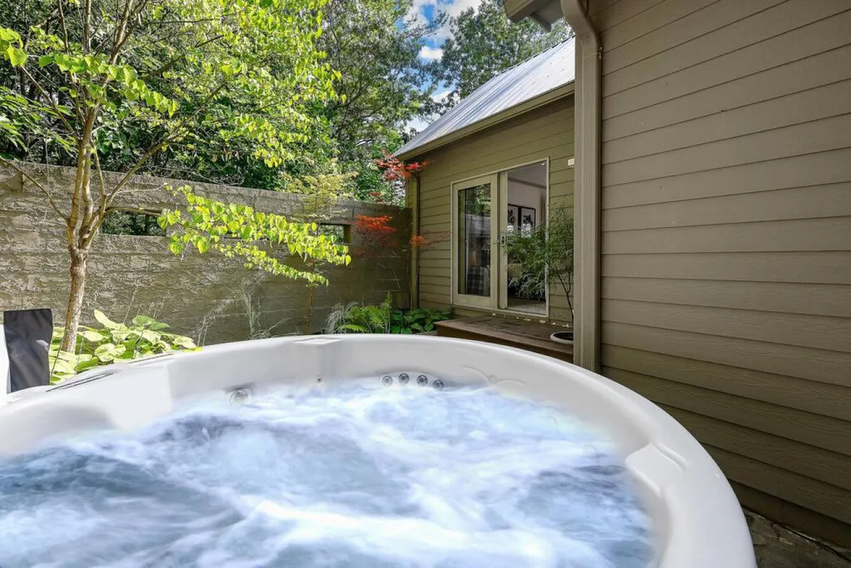 An exterior view of the house featuring a hot tub surrounded by greenery, with inviting windows looking into the home. The scene combines the comfort of home with the serenity of nature, suggesting spaces for fun and relaxation after a day of adventure.
