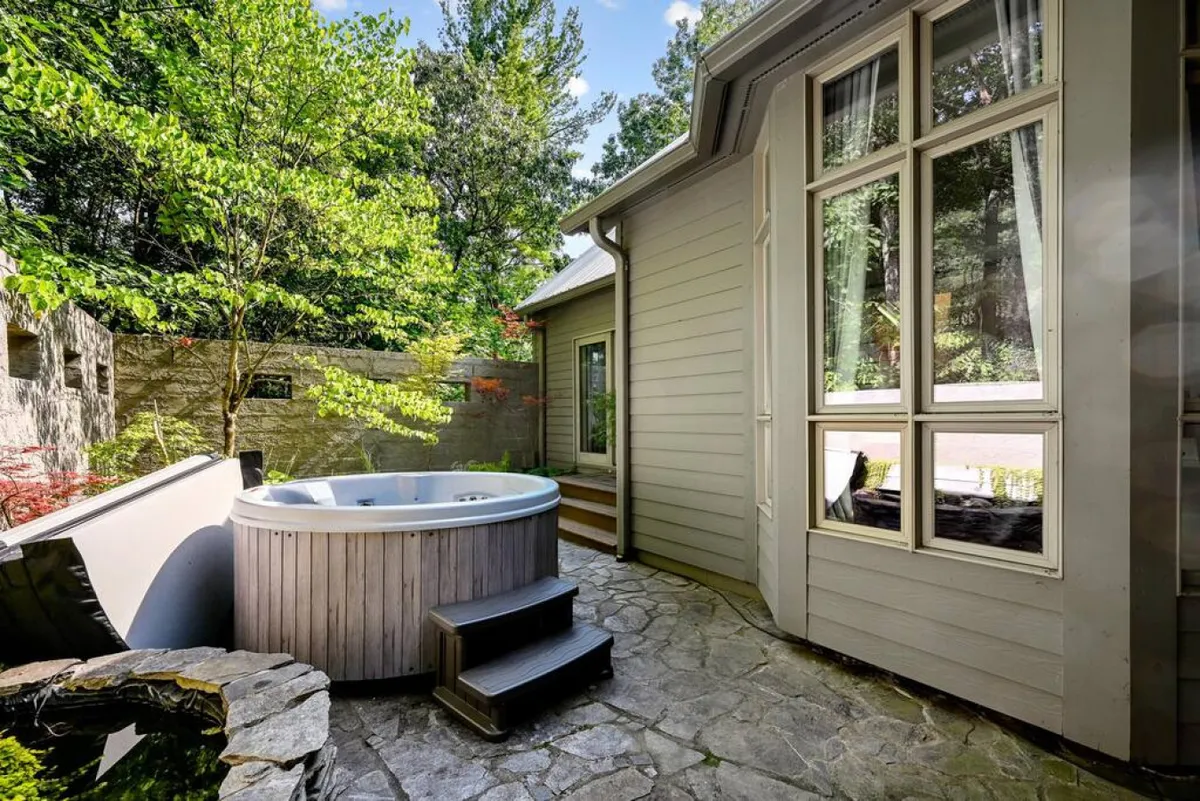This exterior image highlights the inviting 6-person hot tub set amidst lush greenery, adjacent to the home. The stone path and large windows add to the sense of tranquility, while the timing of the photograph captures soft sunset light, suggesting the perfect conditions for evening relaxation or stargazing.