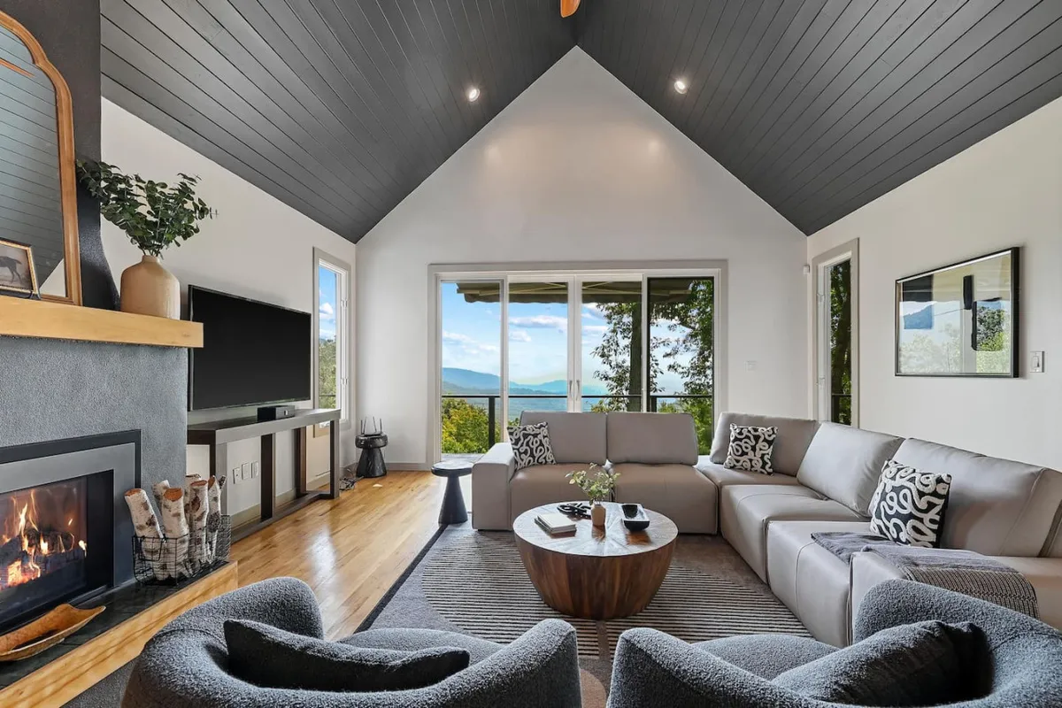 The image showcases a spacious, inviting living area with plush seating arranged around a glowing fireplace. Large windows frame sweeping mountain views, while natural light floods the room, creating a warm and tranquil atmosphere perfect for relaxing or socializing.