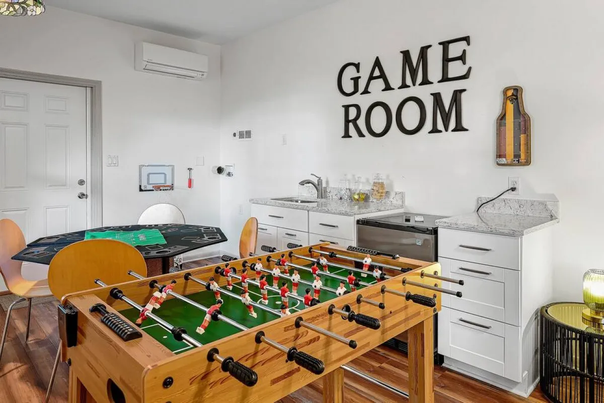 A vibrant game room featuring a foosball table, a dedicated game table, a kitchenette area, and bold wall lettering reading 'GAME ROOM.' The open layout and ample lighting set a lively, social atmosphere perfect for group activities.