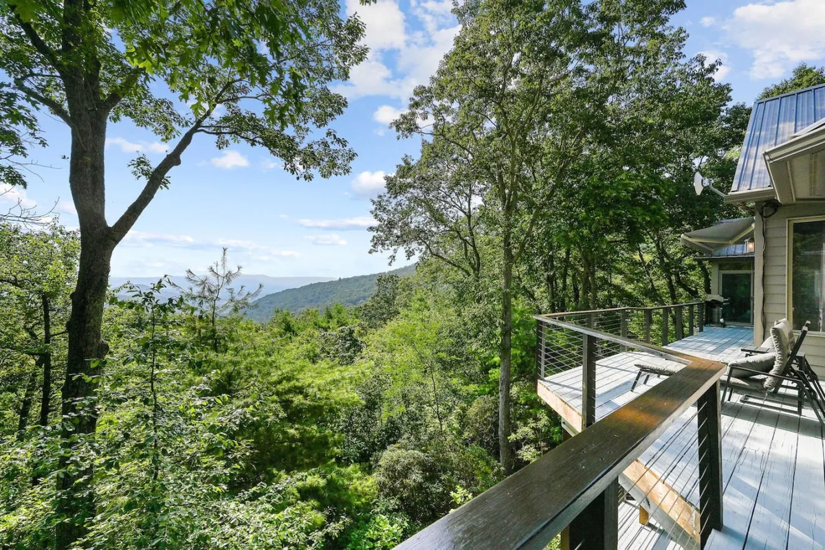 An elevated deck with a classic wooden railing overlooks a sweeping mountain landscape, framed by mature trees. The space is ideal for unwinding, soaking in breathtaking views, or sharing a meal outdoors while surrounded by nature.