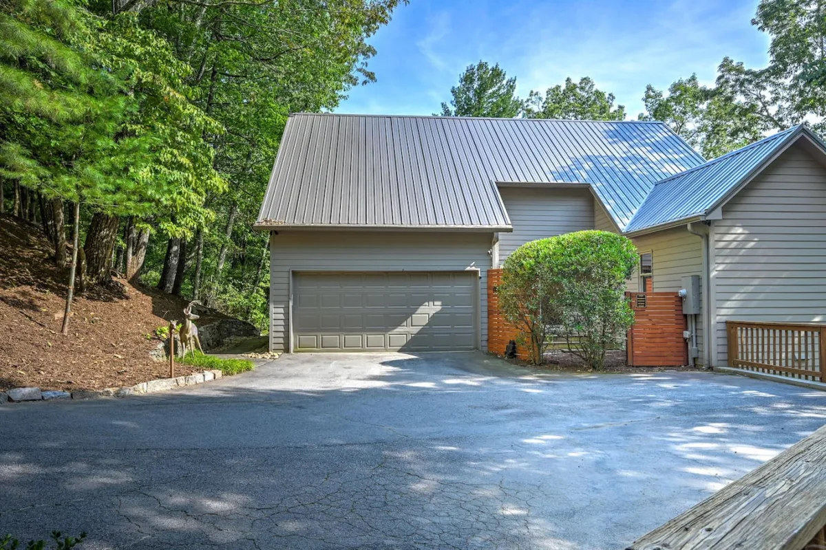 The image shows the home’s attractive exterior with a well-maintained garage door, landscaped shrubbery, a welcoming driveway, and wooden railing. The natural surroundings set a peaceful, upscale tone for arriving guests.