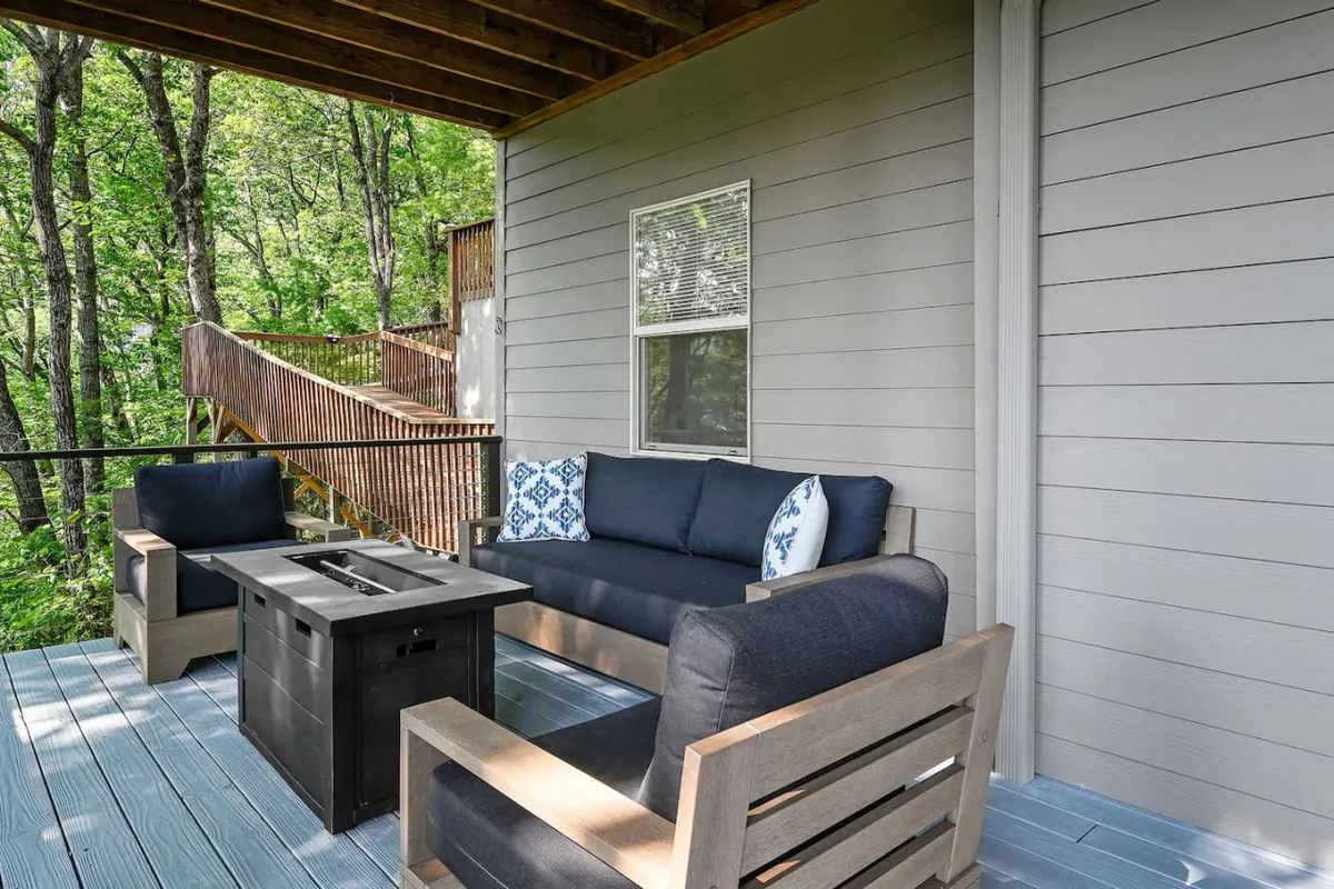 A cozy outdoor seating arrangement encircles a modern fire pit, all set on a wooden deck with abundant surrounding greenery. The space is designed for intimate gatherings, storytelling, and moments of reflection by the warmth of the fire.