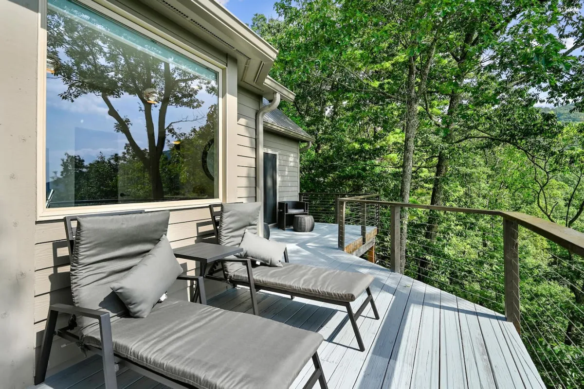 This exterior image features lounge chairs on a wooden deck, bordered by a wooden railing and surrounded by lush trees. A large window offers a glimpse into the interior, and the setting exudes tranquility and connection to the natural landscape.